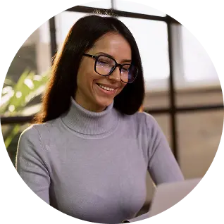 Smiling woman wearing glasses and a light gray turtleneck working on a laptop indoors.