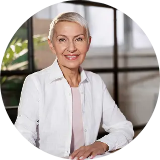 Smiling middle-aged woman with short gray hair wearing a white shirt sitting indoors with large windows in the background.