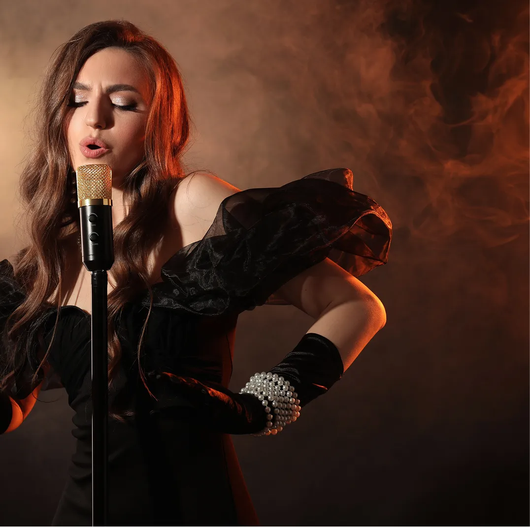 Woman in black dress and gloves singing into a vintage-style microphone with smoky background.