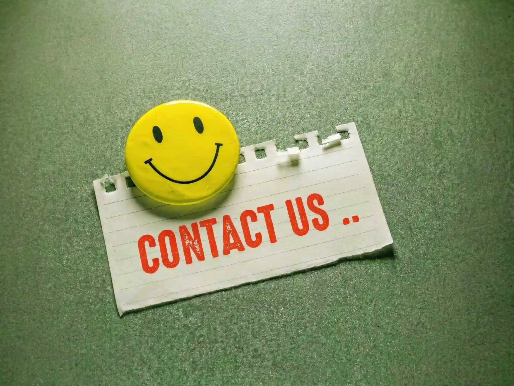 Yellow smiley face button pinned to a torn paper with red text saying 'CONTACT US ..' on a textured green background.