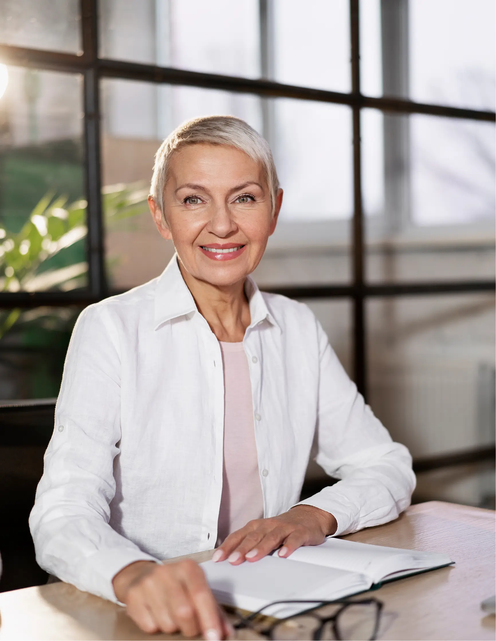 Smiling middle-aged woman with short gray hair sitting at a desk with an open notebook and eyeglasses.