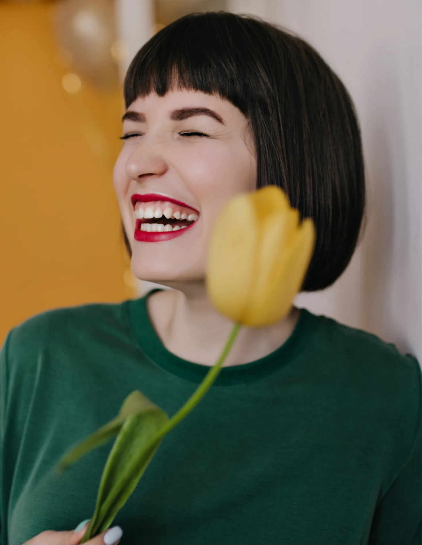Woman with short dark hair and red lipstick laughing and holding a yellow tulip.